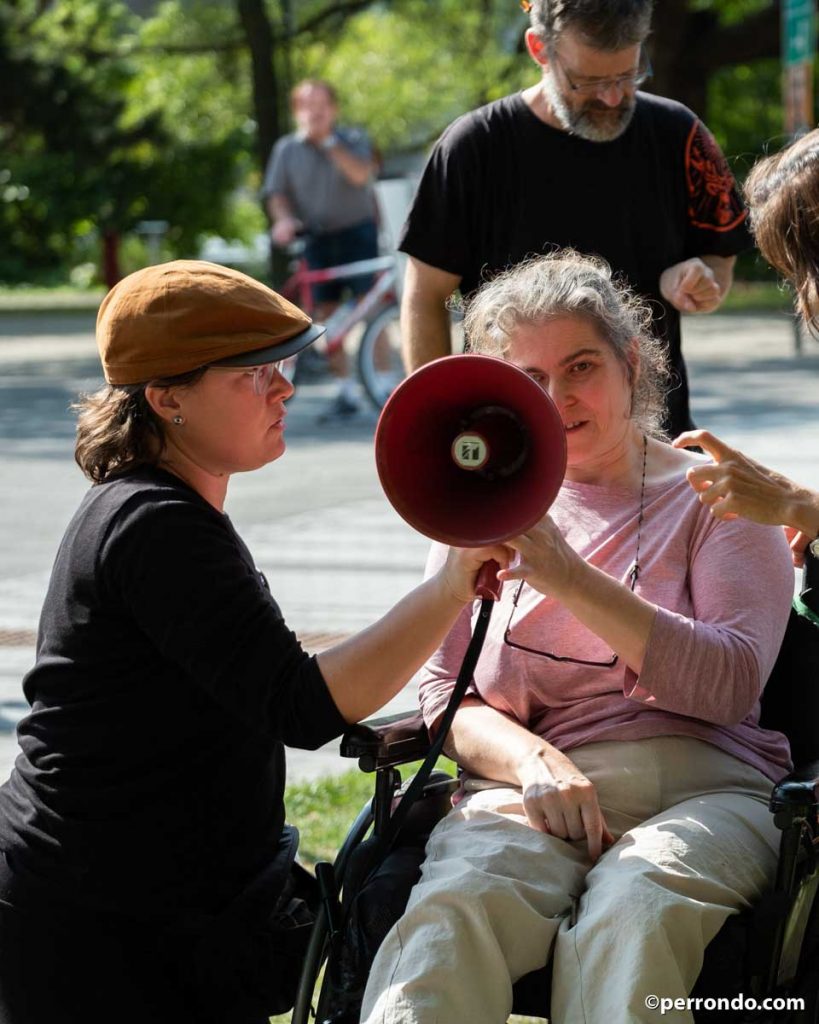 Une femme agenouillée tient un porte-voix rouge pour une femme en fauteuil roulant qui prend la parole.