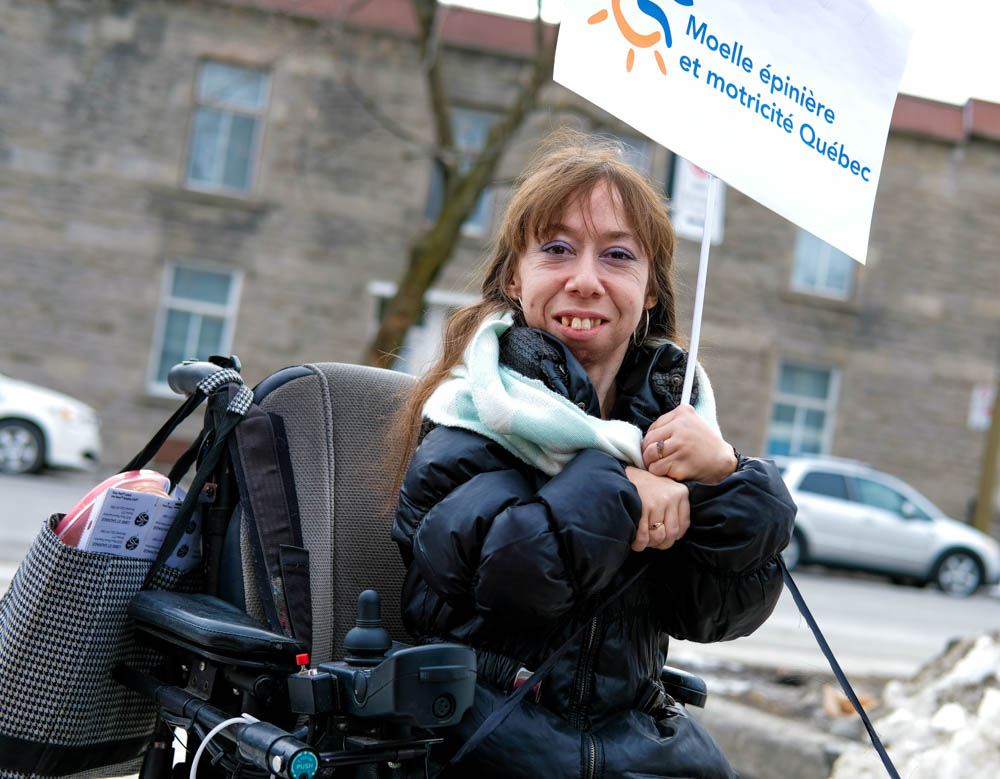 Une femme dans son fauteuil sourit et tient une pancarte sur laquelle on peut lire Moelle épinière et motricité Québec