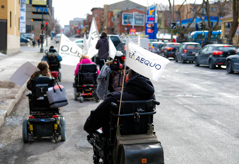 Des personnes en fauteuil roulent dans la rue. Sur leur fauteuil est fixé un fanion blanc sur lequel il est indiqué Ex aequo
