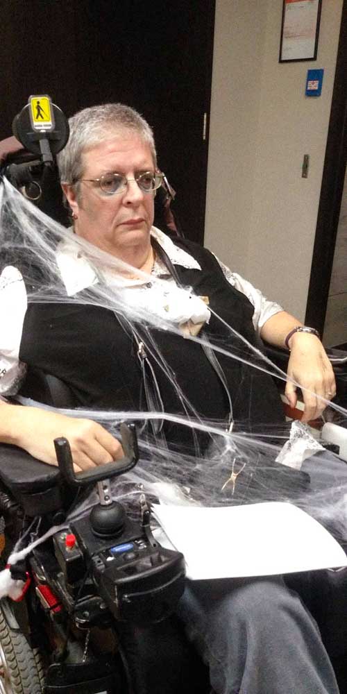 Une femme dans son fauteuil est couverte de toile d'araignée. Elle porte un maquillage sombre et affiche une expression triste.