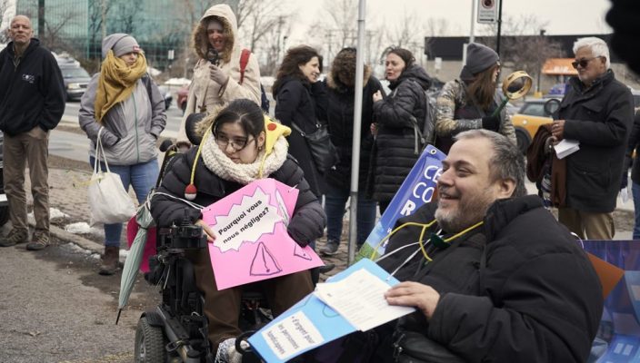 Un groupe de manifestants tenant des pancartes sont réunis. Plusieurs d'entre eux sont en fauteuil roulant.
