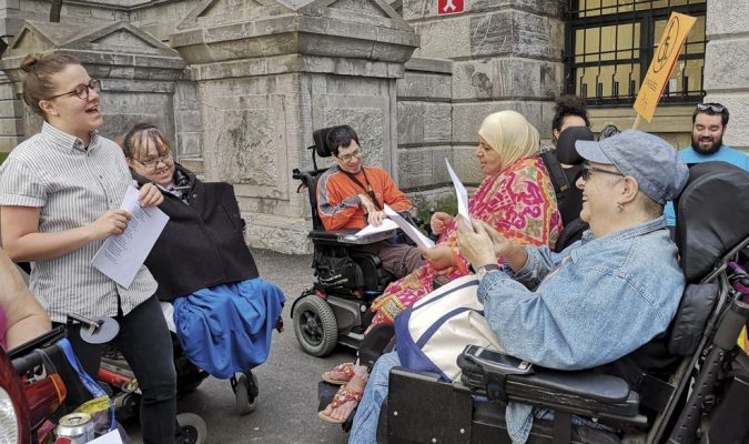 Des personnes en situation de handicap sont rassemblées en cercle devant l'hôtel de ville de Montréal et crient des slogans. 