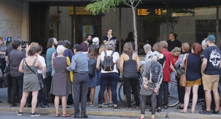 Une foule est rassemblée devant un édicule de métro, de dos. Une quarantaine de personne écoutent la lecture d'un texte par une femme au porte-voix.