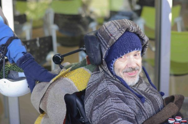 Un homme en fauteuil roulant sourit à la caméra. Il porte une tuque et un manteau très chaud.